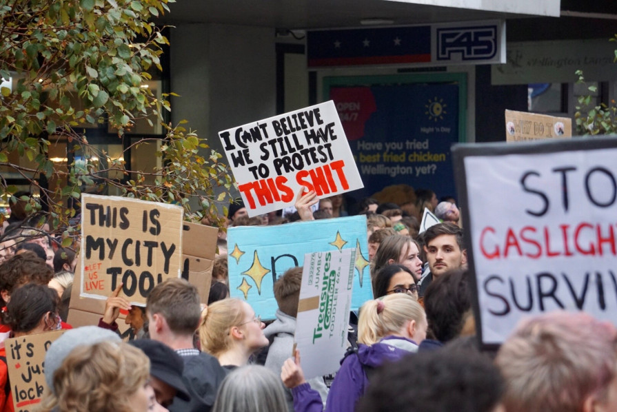 A group protest, with someone holding up a sign saying "I can't believe we still have to put up with this shit"